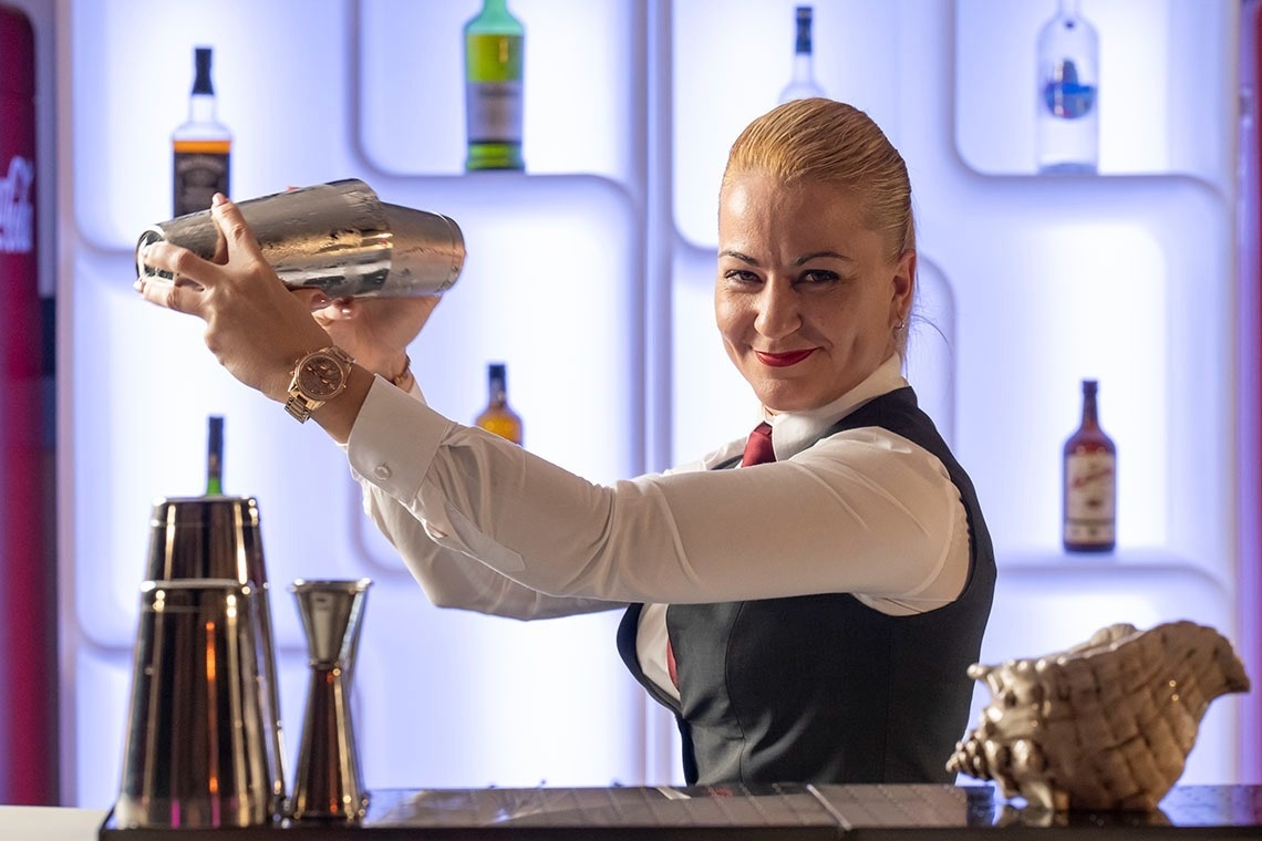 Una mujer camarera sonríe mientras agita una coctelera en un bar con botellas iluminadas de fondo.