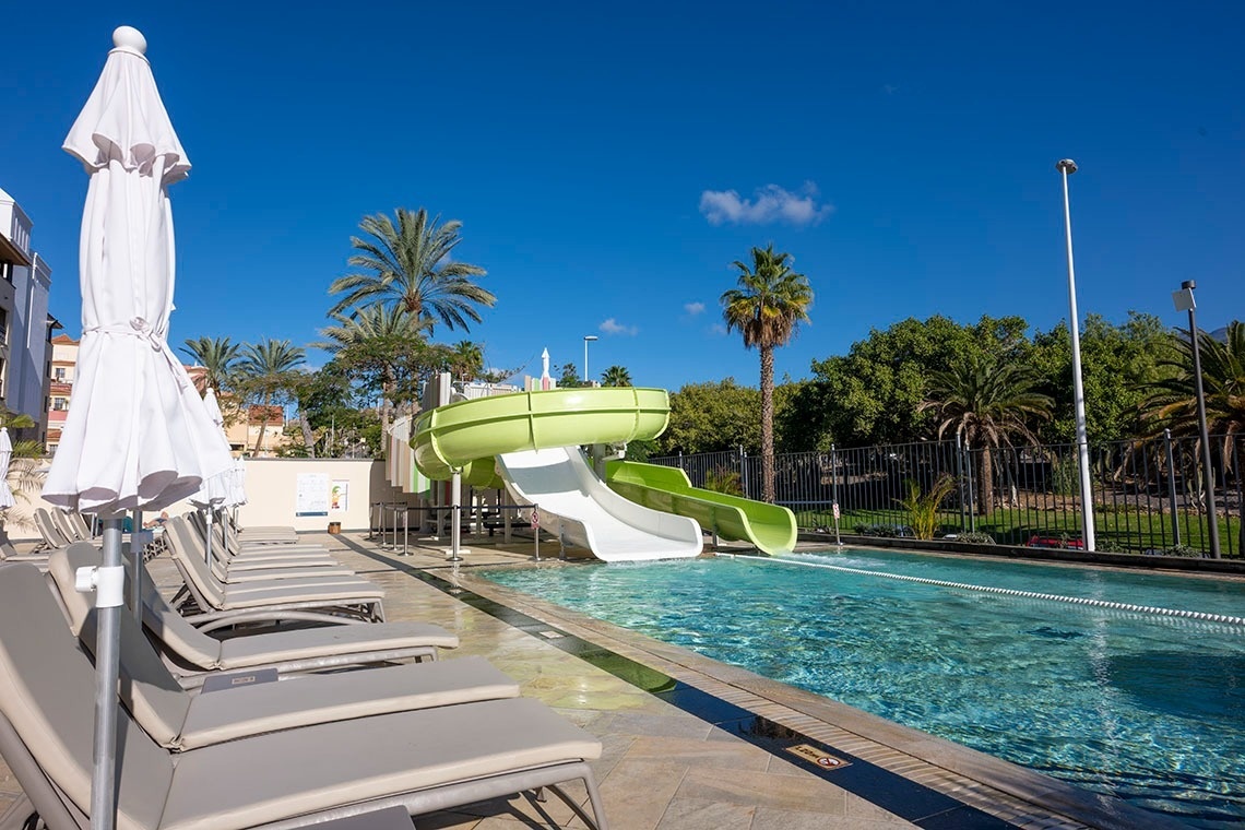 Una piscina exterior con toboganes de agua verdes, hamacas blancas y palmeras bajo un cielo azul claro.
