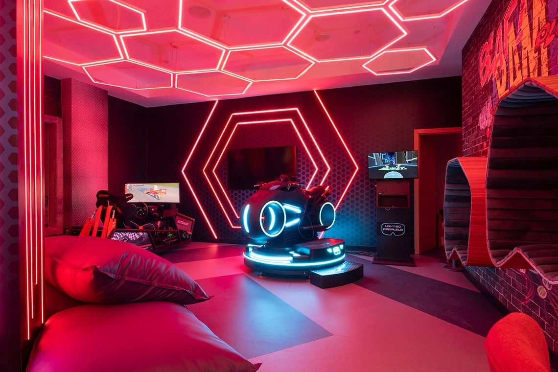 A vibrant, futuristic gaming room is illuminated by red and blue neon lights, featuring hexagonal patterns, racing simulators, a central motion ride, and modern seating areas.