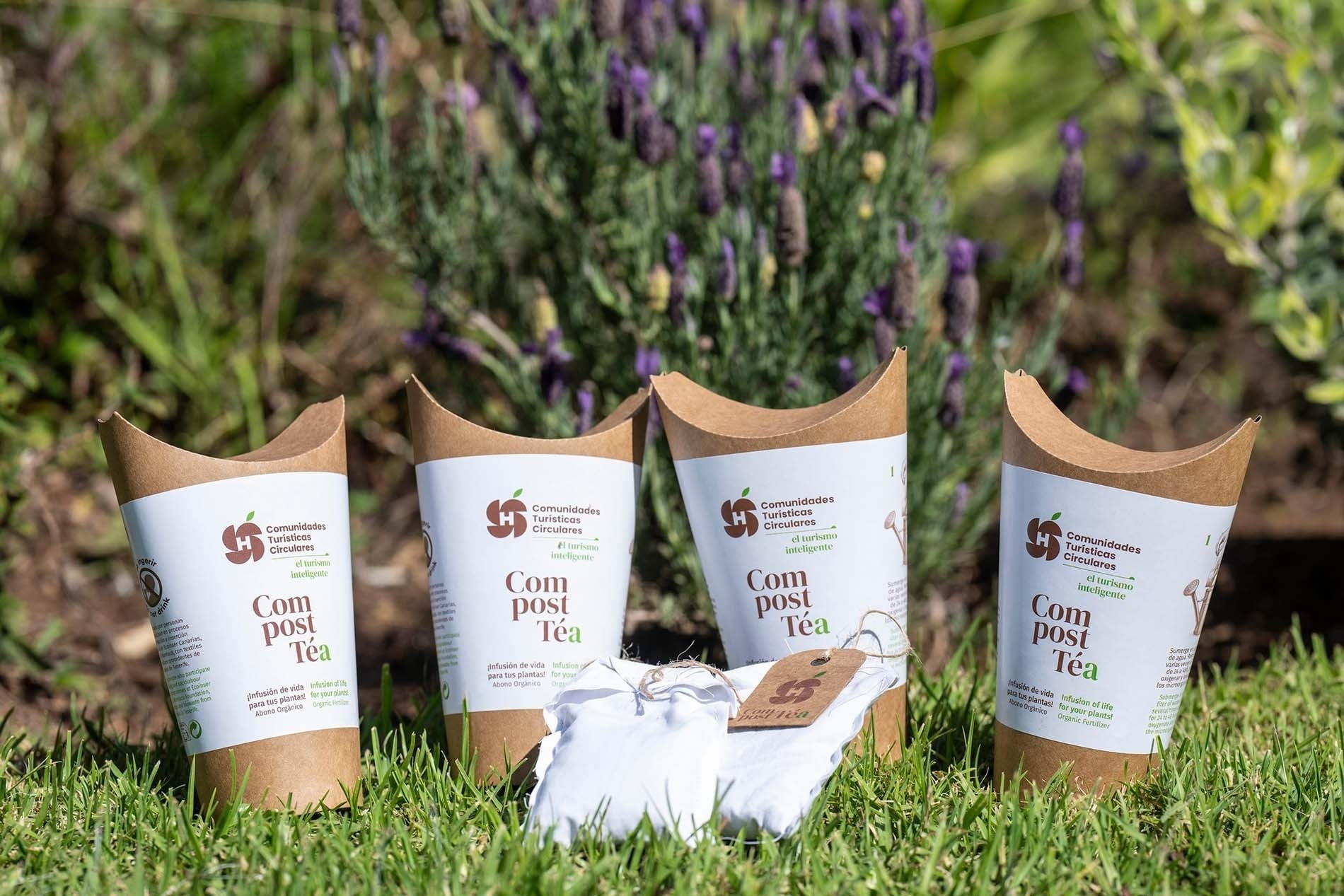 Cuatro envases de "Compost Téa" y dos pequeñas bolsas blancas reposan sobre césped verde con lavanda borrosa al fondo.