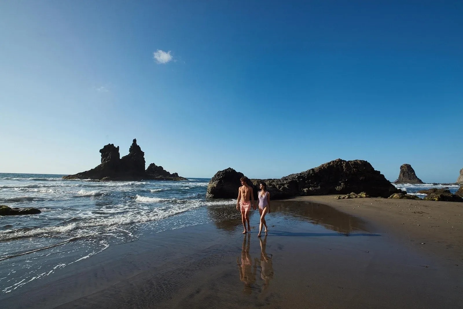 un hombre y una mujer caminan por la playa con una gran roca en el fondo