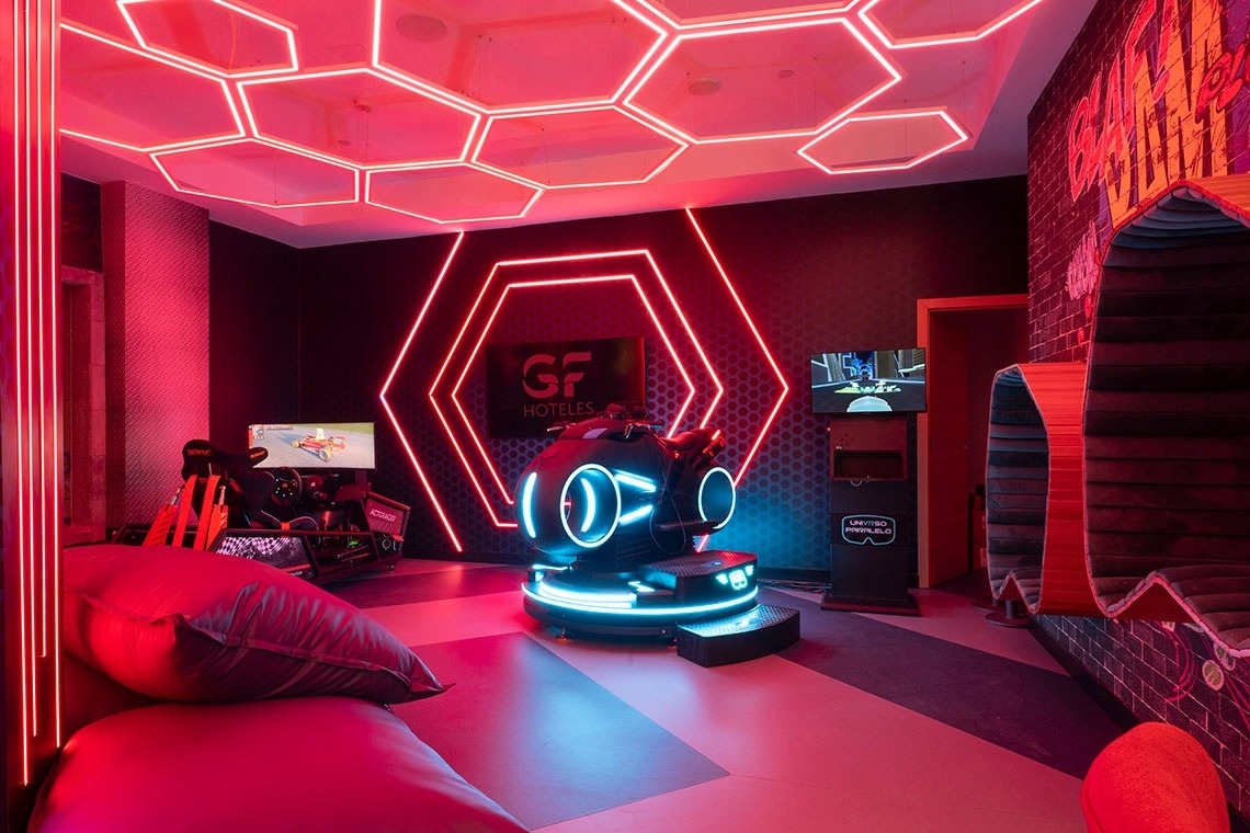 A vibrant gaming room features red neon hexagonal lights on the ceiling and walls, a racing simulator, a futuristic glowing blue gaming pod, and various seating options.