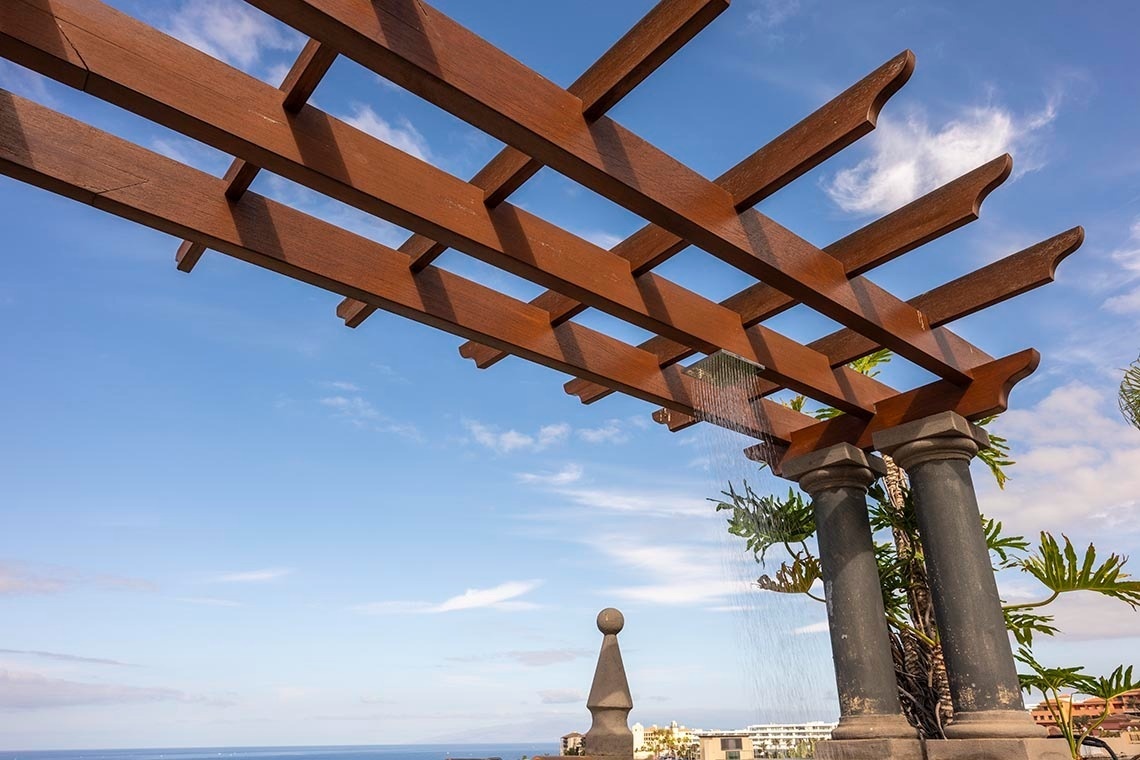 An outdoor shower under a wooden pergola, supported by dark stone pillars, is set against a bright blue sky with a distant view of the ocean and buildings.