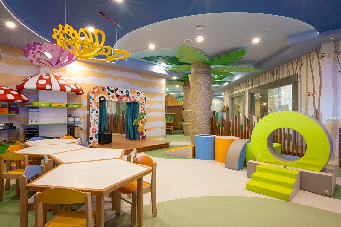 This image captures a vibrant and whimsical children's play area, featuring colorful furniture, nature-themed decor like mushroom shelves and palm tree structures, a puppet stage, and soft play equipment.