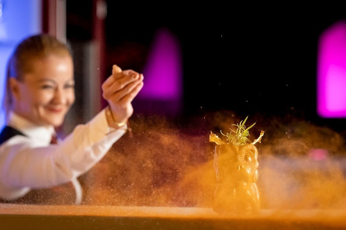 Una mujer sonriente rocía polvo naranja alrededor de un cóctel decorado en un vaso con forma de cabeza, creando un efecto visual vibrante.