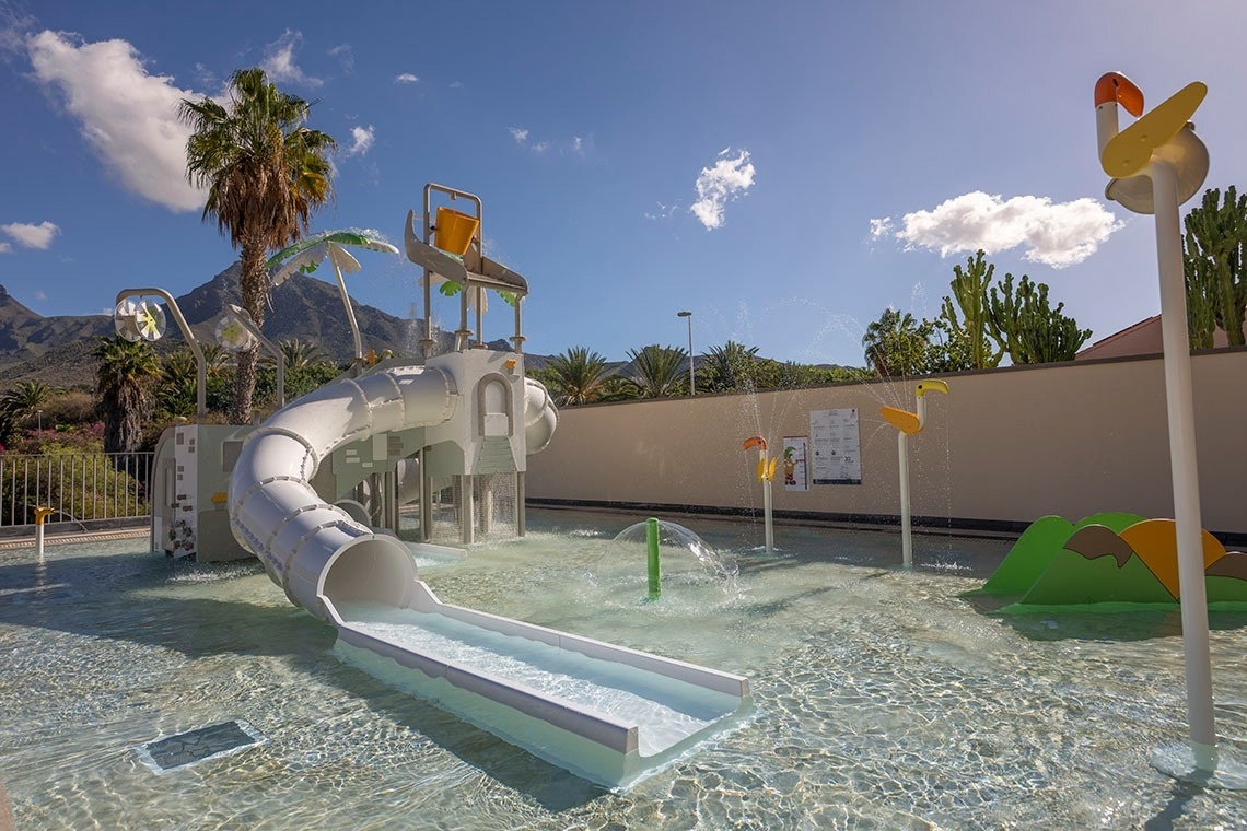 Una soleada zona de juegos acuáticos para niños con un tobogán blanco, chorros de agua y elementos lúdicos, enmarcada por montañas y palmeras.