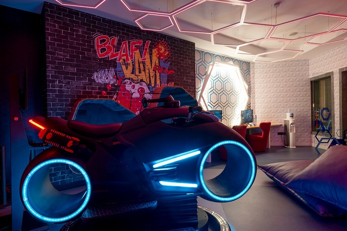 A futuristic light cycle with blue neon lights sits prominently in a gaming-themed room adorned with graffiti, hexagonal ceiling lights, and geometric patterned walls.