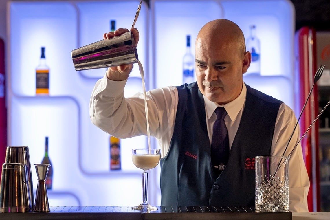 Un camarero calvo, vestido con camisa blanca, chaleco oscuro y corbata, vierte cuidadosamente una bebida blanca de una coctelera a una copa, con botellas iluminadas en el fondo.