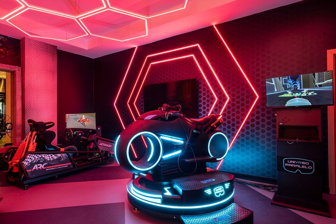 A futuristic, neon-lit gaming arcade features a Tron-style motorcycle VR simulator and a racing simulator, set amidst a dark room with glowing red hexagonal patterns on the ceiling and walls.