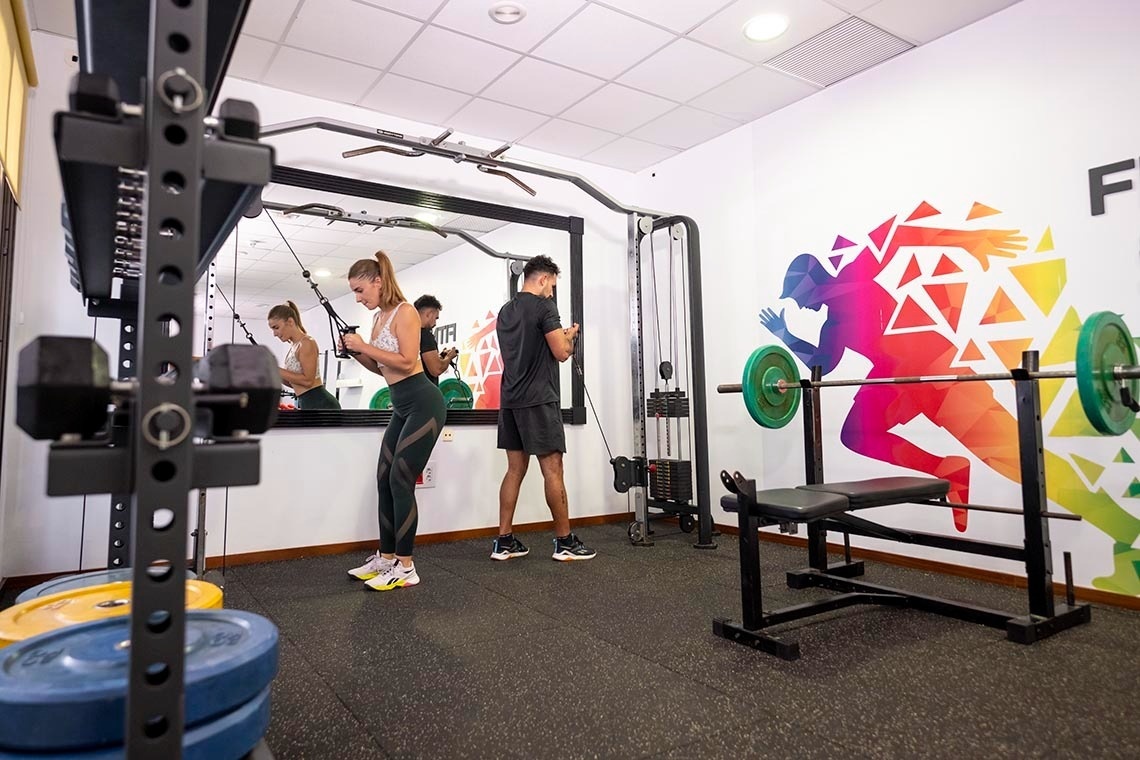 Two people are exercising in a gym with various fitness equipment, including a squat rack, cable machine, and bench press, alongside a colorful wall mural of a runner.