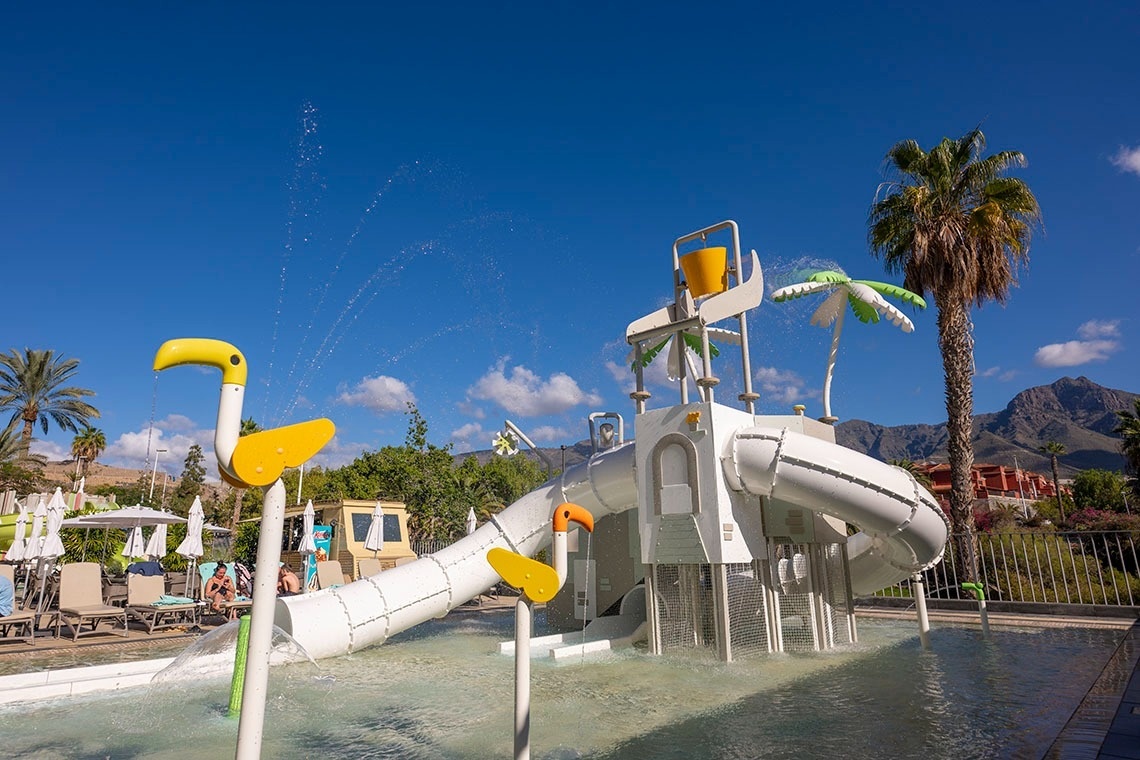 Una escena de un parque acuático con toboganes y juegos de agua, palmeras y montañas bajo un cielo azul y soleado.