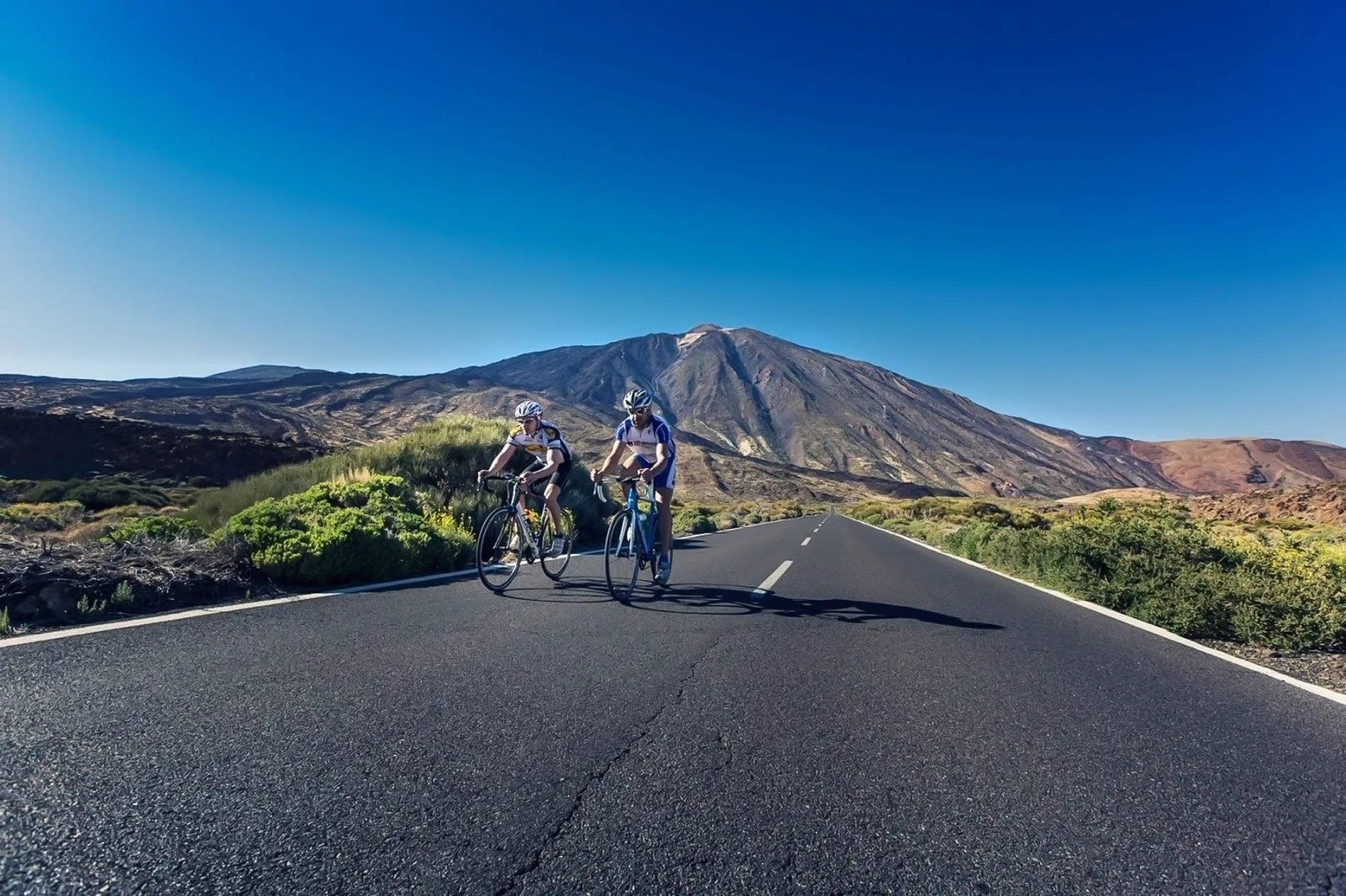 two people riding bicycles down a road with a mountain in the background