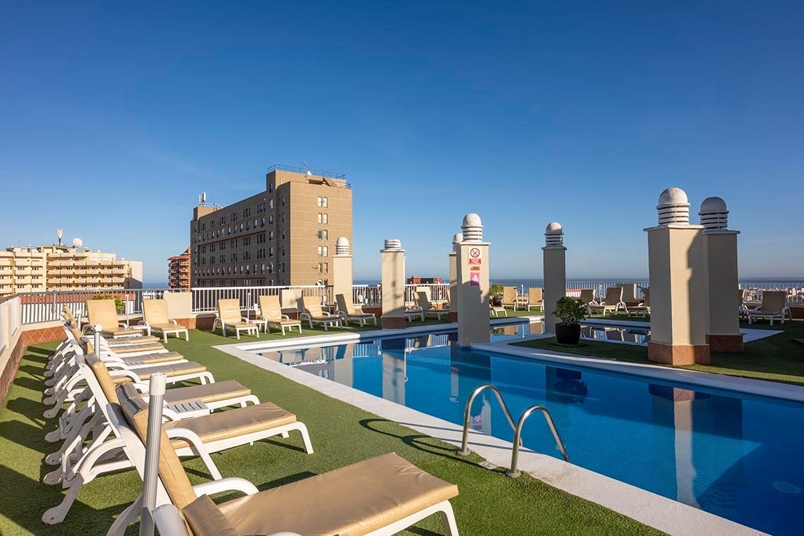 A sunny rooftop swimming pool with lounge chairs sits atop a building, overlooking other structures and the distant ocean under a clear blue sky.