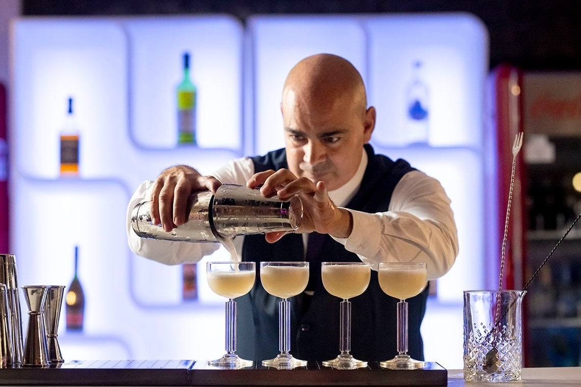 Un barman calvo vierte un cóctel de una coctelera en una fila de copas sobre la barra de un bar iluminado.