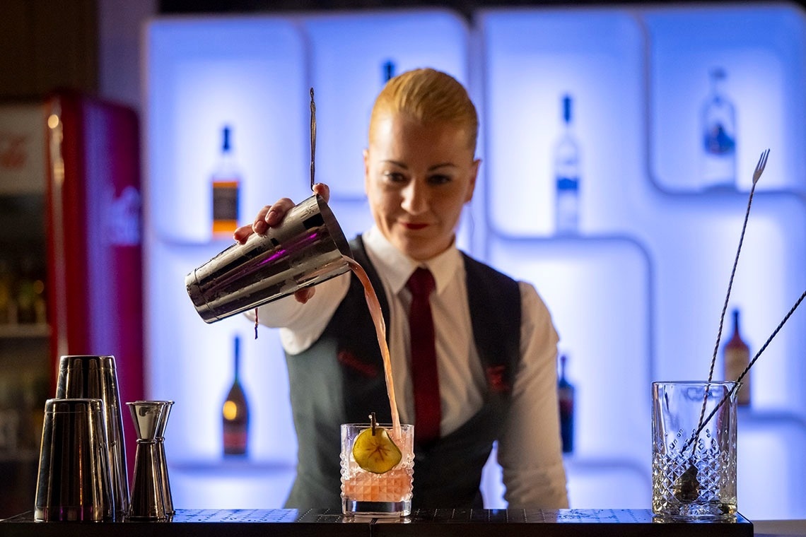 Una mujer barman vierte un cóctel rosado de una coctelera en un vaso adornado, con un fondo de botellas iluminadas en azul.