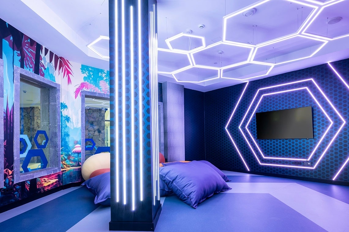 This is a vibrant, modern room featuring hexagonal LED lights on the ceiling and a wall, a tropical-patterned wall, and comfortable floor cushions.
