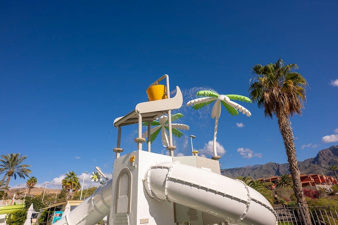 Una zona de juegos acuáticos con toboganes, un cubo volquete amarillo y aspersores con forma de palmera, se encuentra bajo un cielo azul claro con palmeras y montañas al fondo.