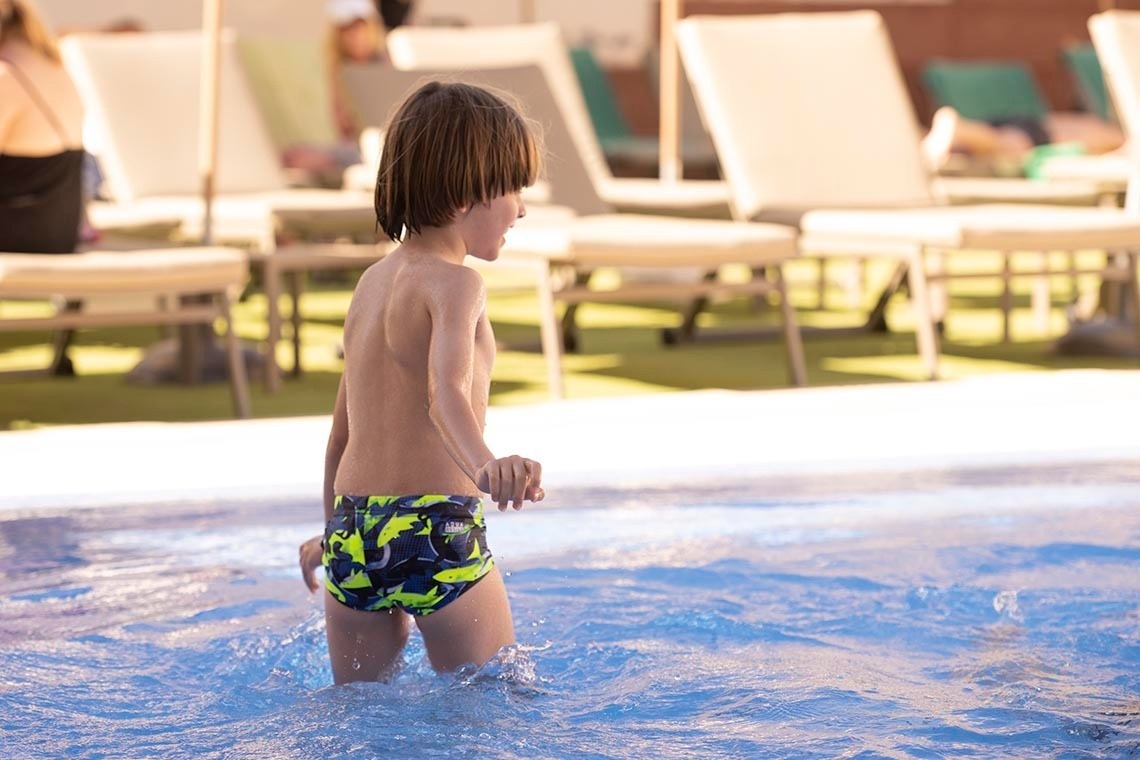 Un niño con bañador de estampado de tiburones está de pie en el agua de una piscina, con tumbonas y personas desenfocadas al fondo.