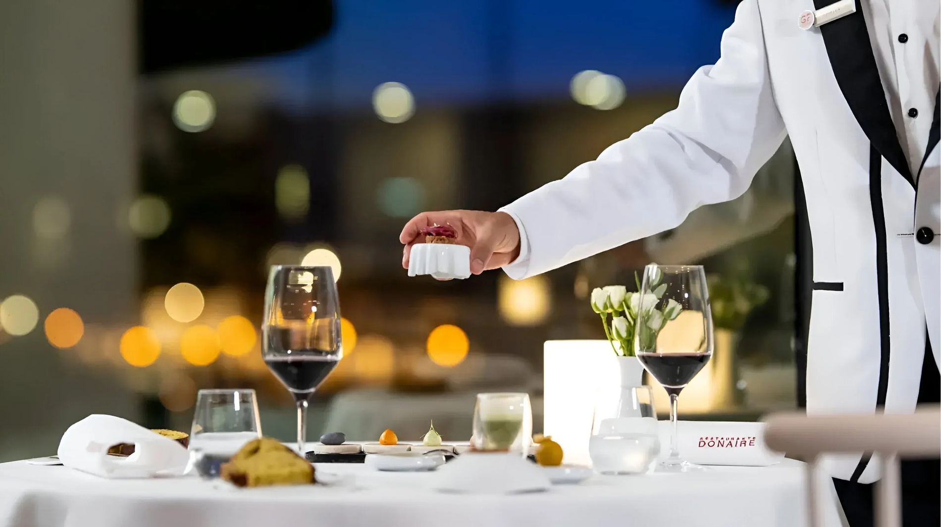 un hombre en una chaqueta blanca sostiene un plato frente a una mesa con copas de vino