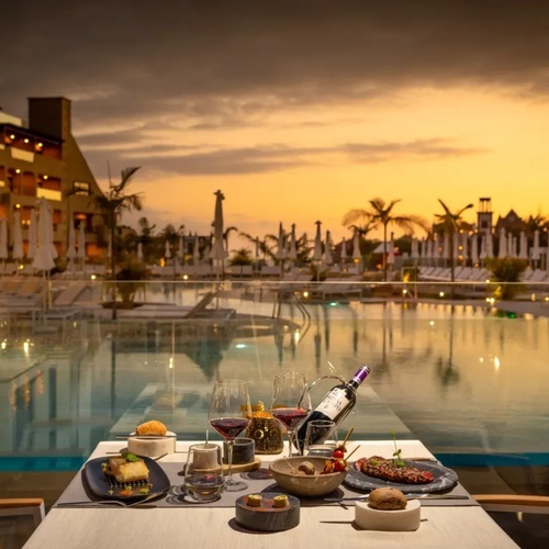 una mesa con comida y una botella de vino frente a una piscina al atardecer