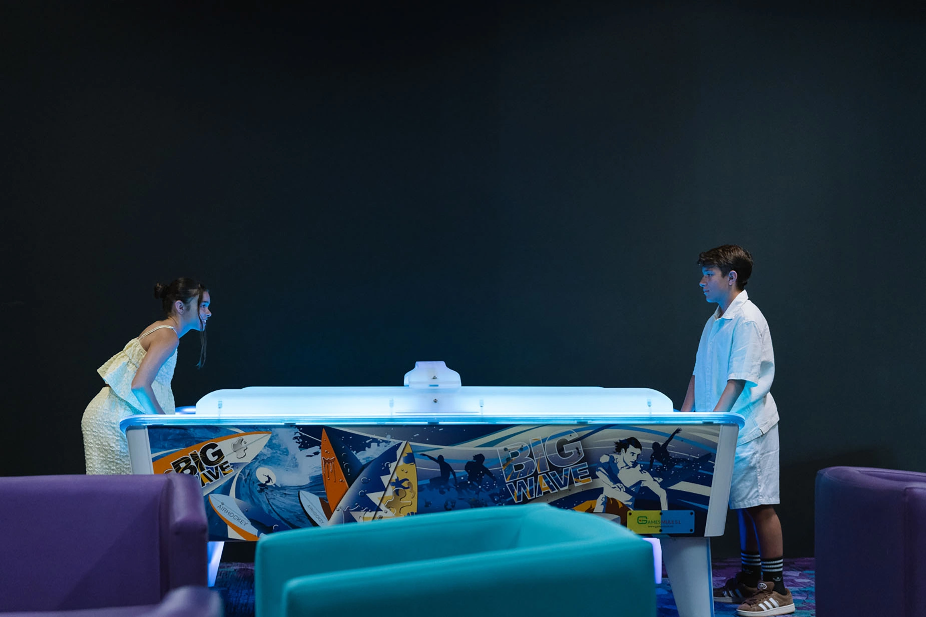 Pareja jugando air hockey en un hotel, con sofás morados y turques. GF: Victoria