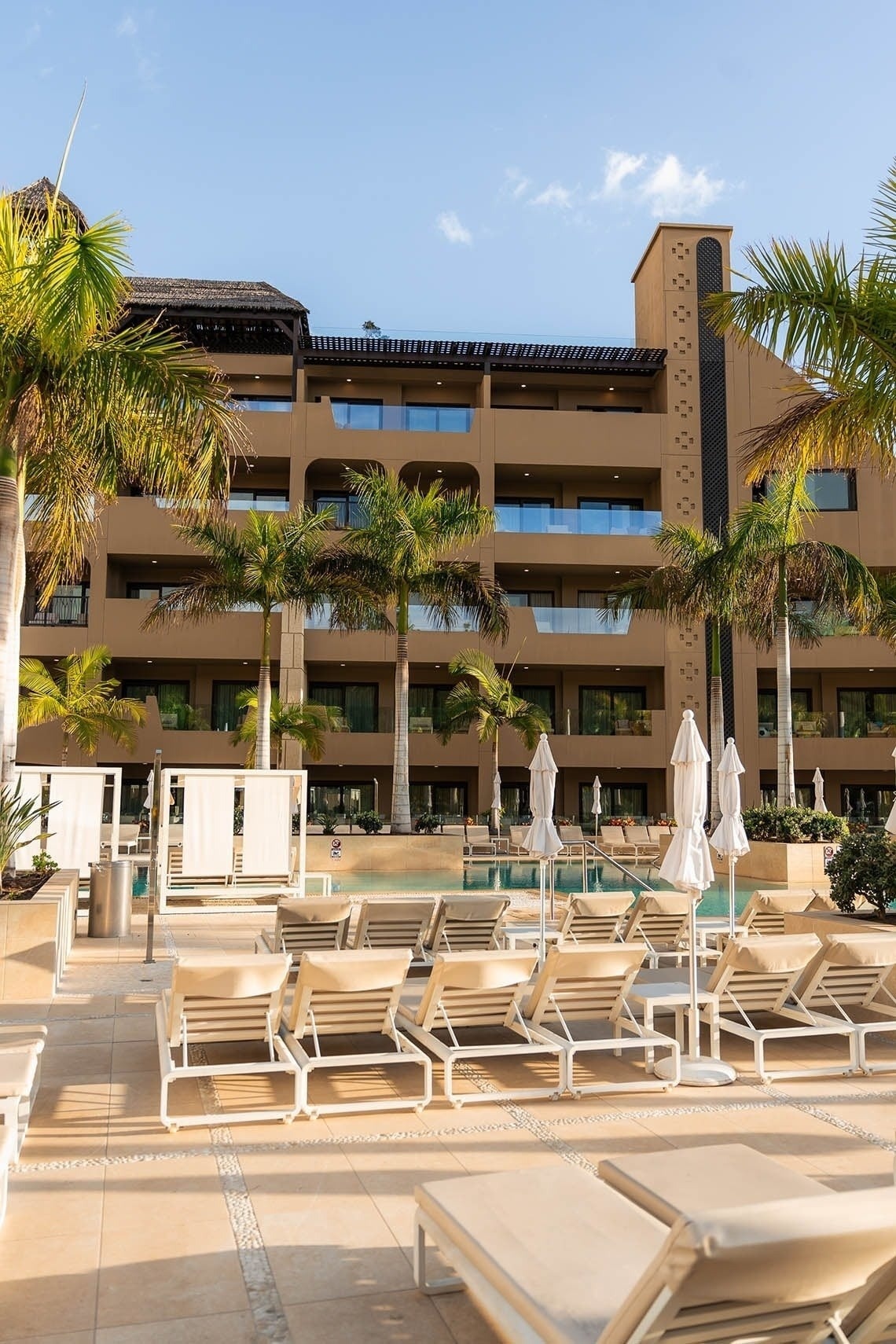 This vibrant resort scene captures a multi-story hotel building surrounded by palm trees, a swimming pool, and numerous lounge chairs under a clear blue sky.