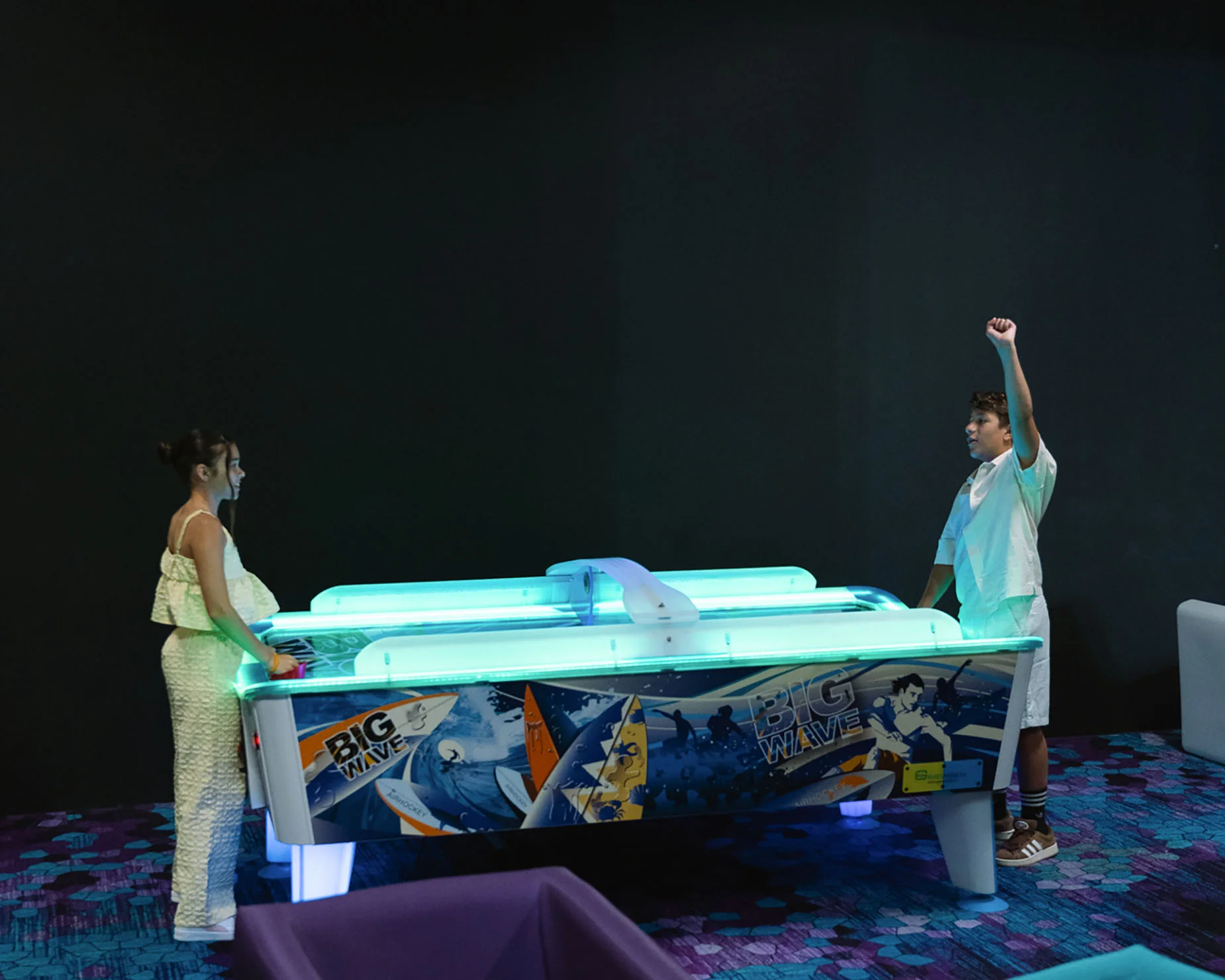 Jóvenes jugando air hockey en la sala de juegos del hotel, GF: Victoria con iluminación LED vibrante.