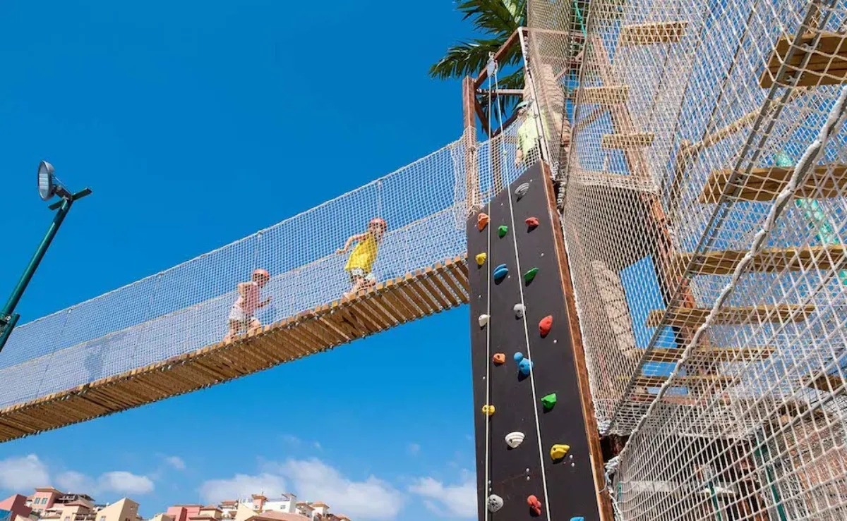 two children are walking across a wooden bridge over a climbing wall