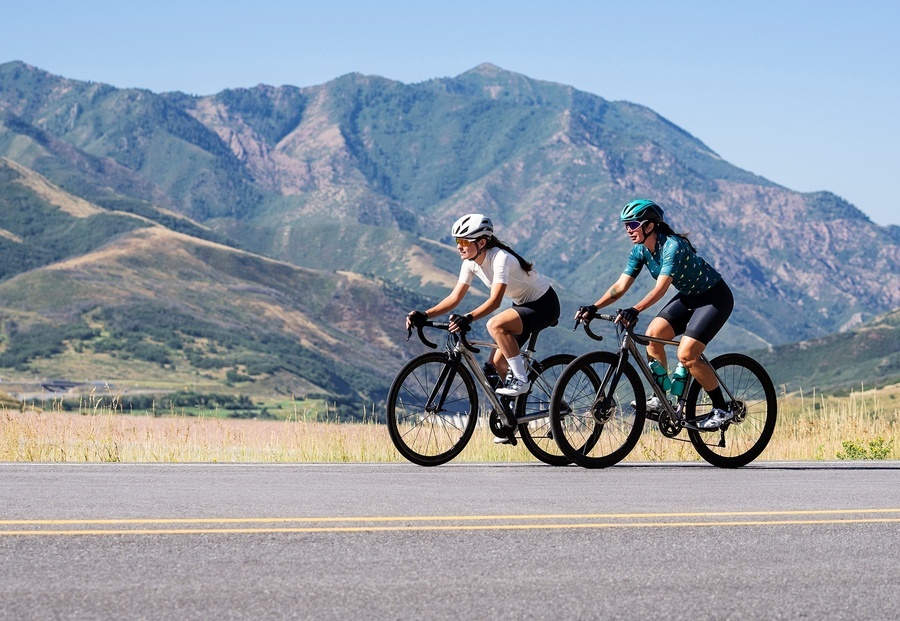 two women riding bicycles down a road with mountains in the background