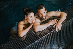 Una pareja sonriente y relajada disfruta en una piscina o jacuzzi, apoyados en el borde de azulejos oscuros.