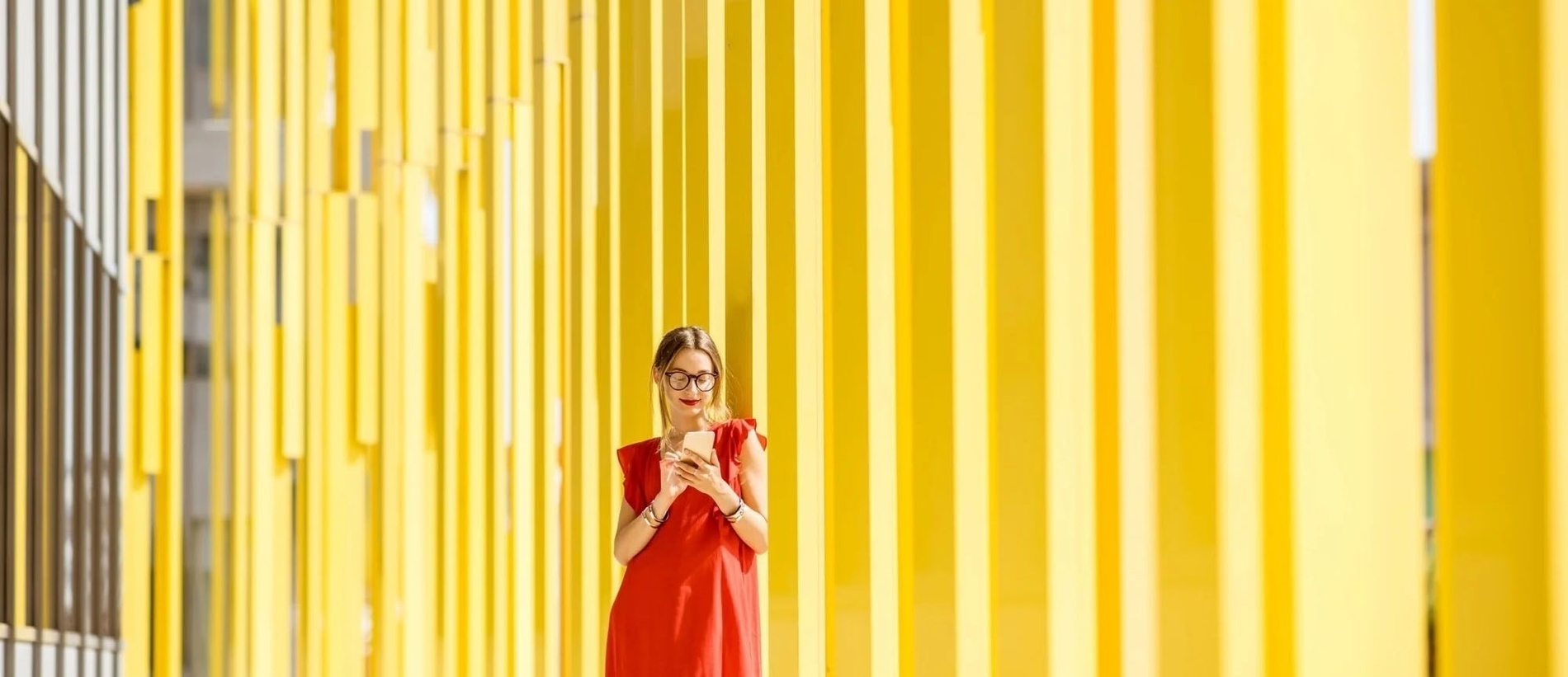 una mujer con un vestido rojo y gafas está parada frente a una pared amarilla y mirando su teléfono .