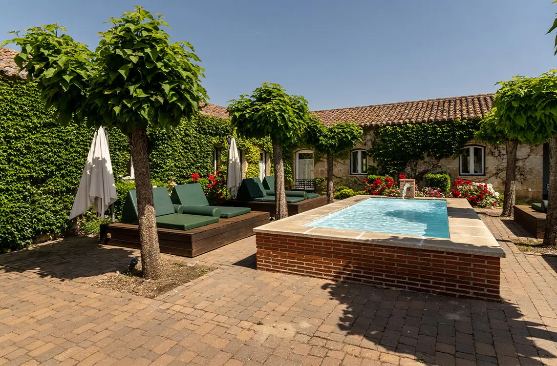 Piscina con fuente y tumbonas verdes en un patio adoquinado, rodeado de vegetación y arquitectura rural. Ideal paraHacienda Zorita Wine Hotel.