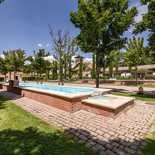 Fuente de ladrillo y piedra con piscina de agua azul, césped verde y árboles en Hacienda Zorita Wine Hotel