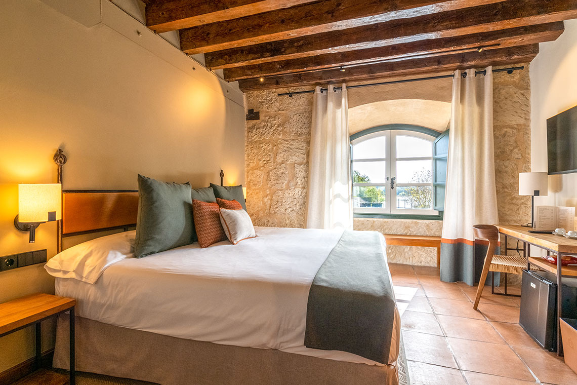 Habitación de hotel rústica con cama king size, vigas de madera y ventana arqueada. Hacienda Zorita Wine Hotel.