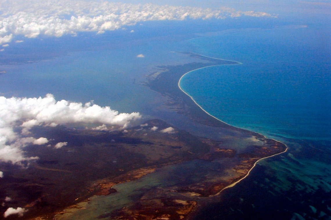 Una vista aérea muestra una extensa y sinuosa línea costera, con aguas oceánicas de distintas tonalidades de azul y nubes sobre el paisaje.