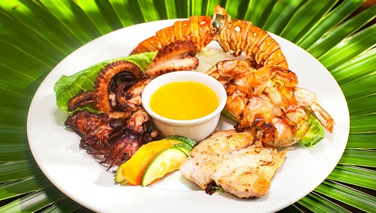 Un plato de mariscos variados a la parrilla, que incluye langosta, pulpo, calamares, camarones y pescado, se presenta con una salsa y vegetales sobre hojas de palma verdes.