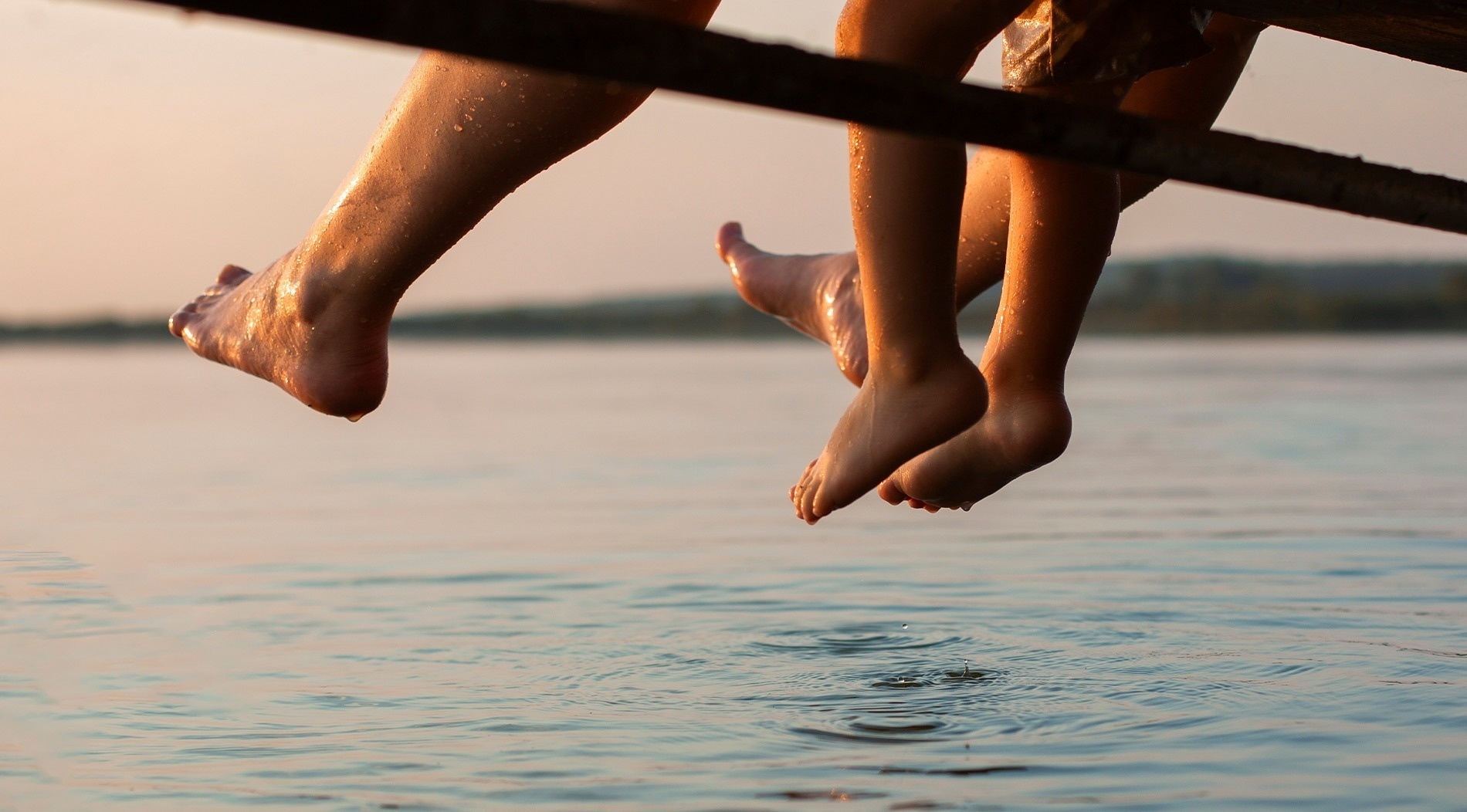 Pies mojados, iluminados por la luz dorada del atardecer, cuelgan sobre el agua tranquila, creando suaves ondas en su superficie.