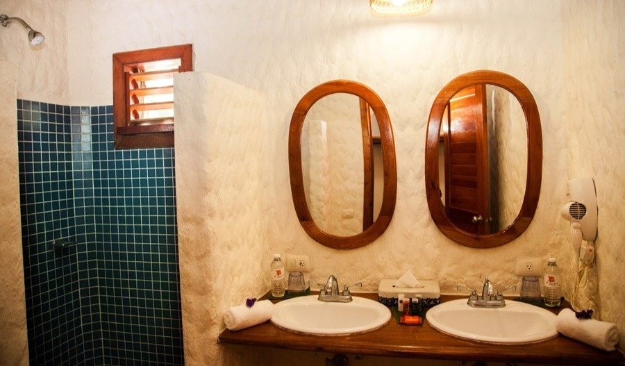 Un baño de estilo rústico que presenta una ducha con azulejos azules, una ventana de madera y un tocador doble con lavabos y espejos ovalados de marco de madera.
