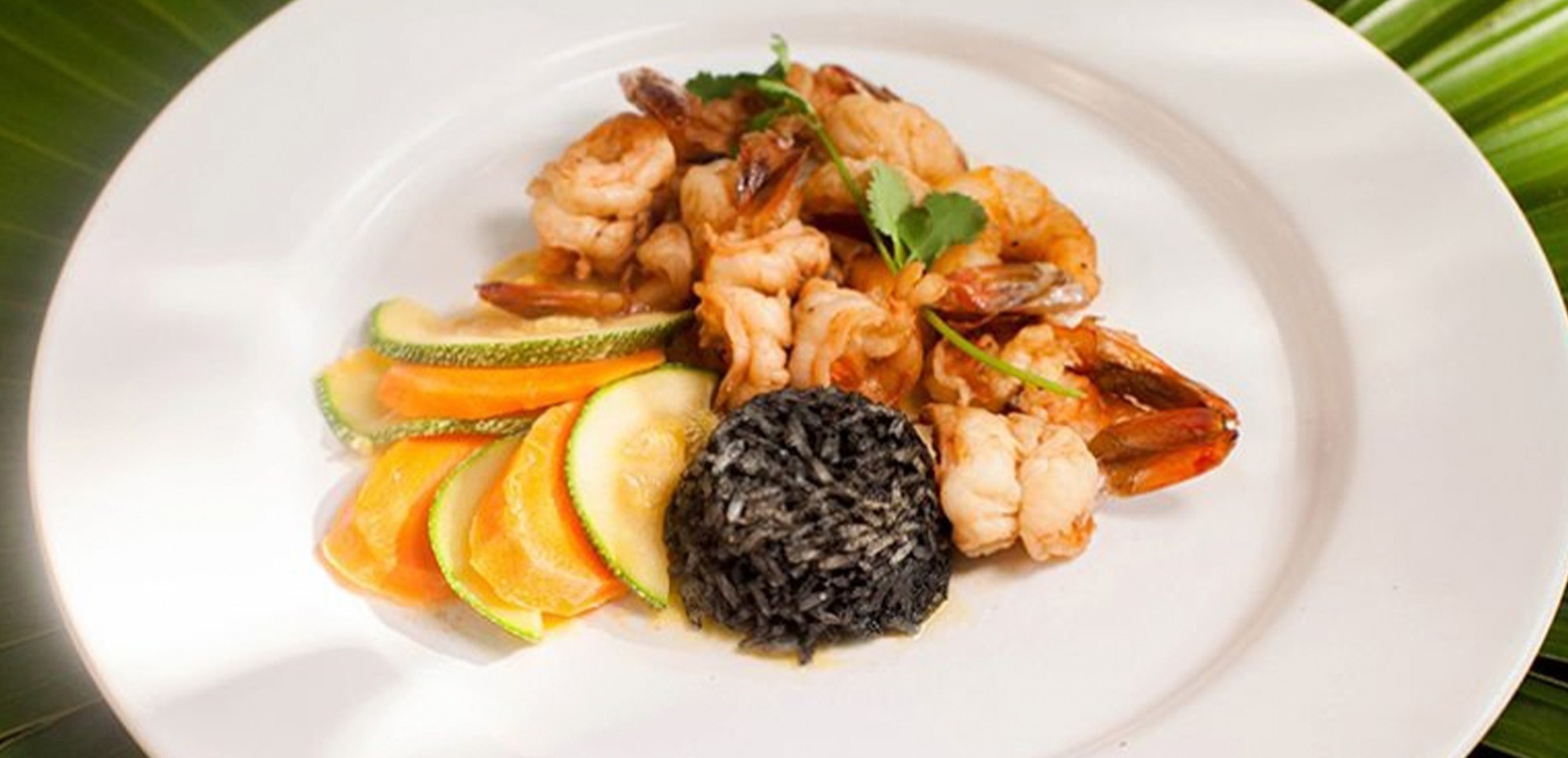 En un plato blanco se aprecian camarones salteados, una porción de arroz negro y rodajas de calabacín y zanahoria, con un fondo de hojas verdes.
