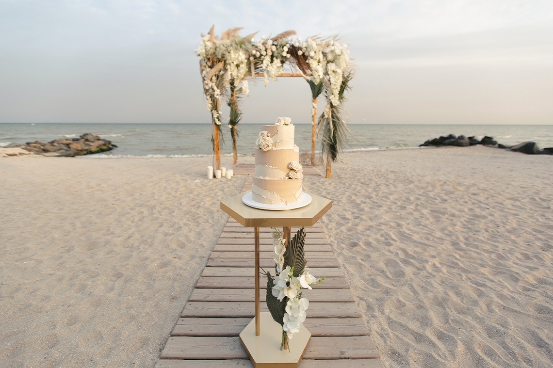 Un pastel de bodas de tres niveles se exhibe en un pedestal sobre una pasarela de madera en una playa, con un arco floral decorado al fondo.