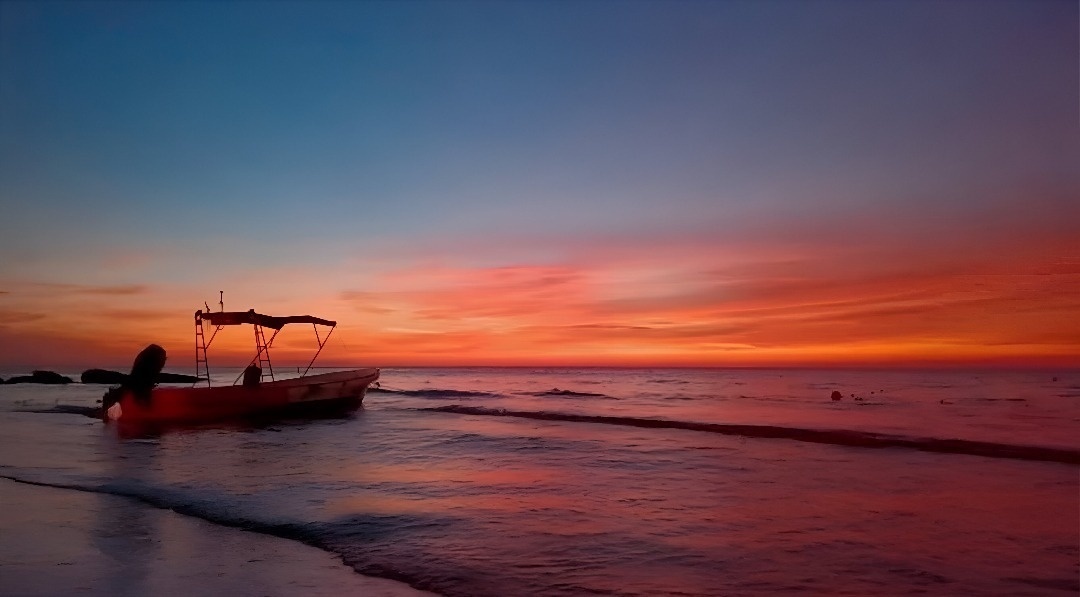 Un bote descansa en la orilla de una playa al atardecer, con el cielo y el mar teñidos de vibrantes azules, naranjas y rojos.