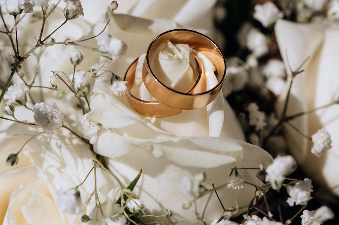 Dos anillos de boda de oro descansan sobre una rosa blanca, rodeados de pequeñas flores blancas.