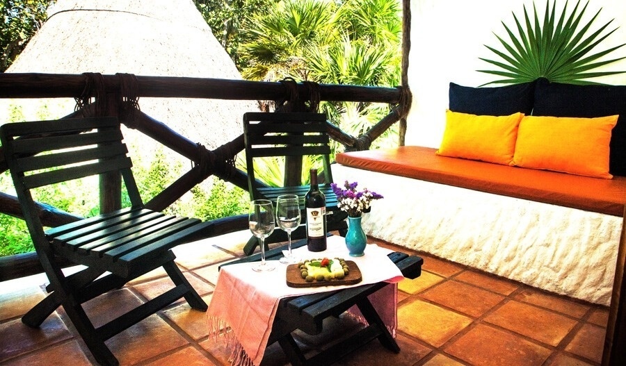 Acogedor balcón tropical con sillas y mesa de madera, vino y aperitivos, un banco con cojines naranjas y exuberante vegetación de fondo.