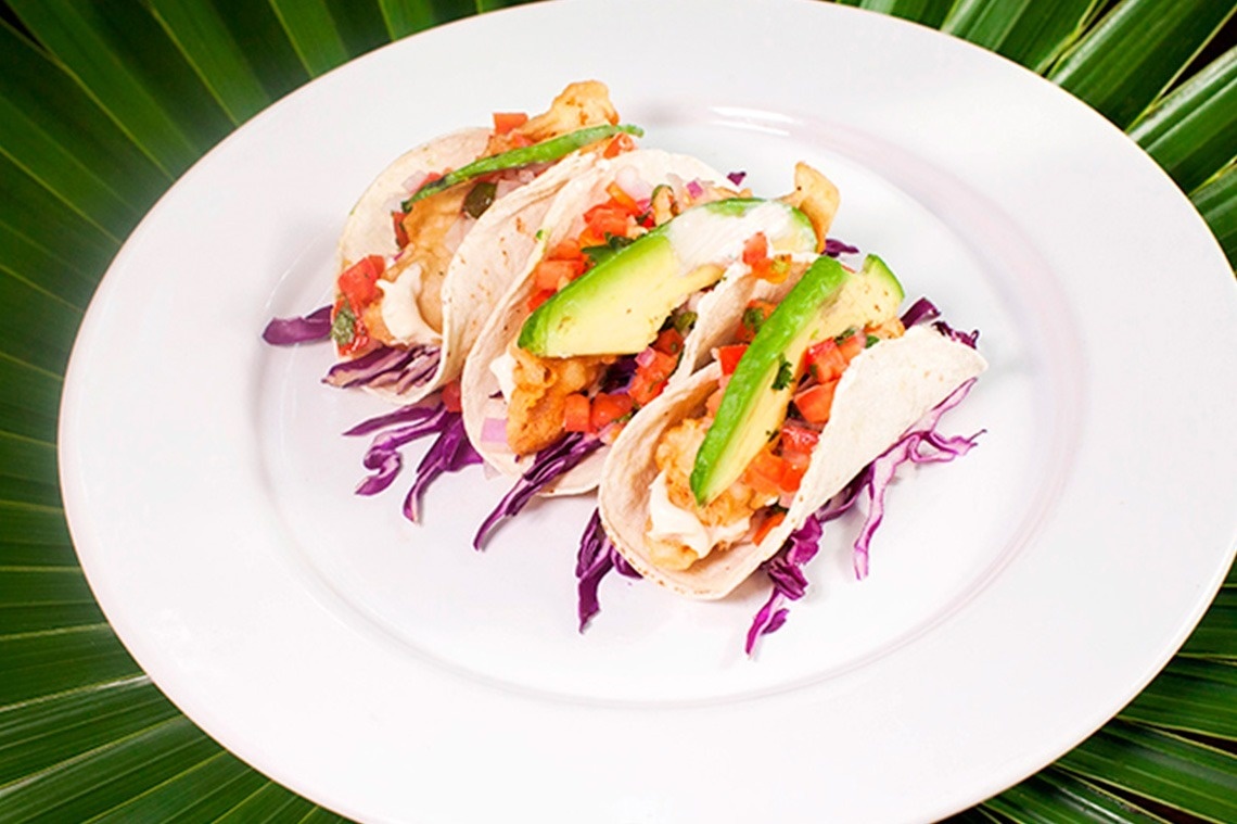 Tres tacos con pescado frito, pico de gallo, aguacate y col morada se presentan en un plato blanco sobre un fondo de hojas de palma verdes.