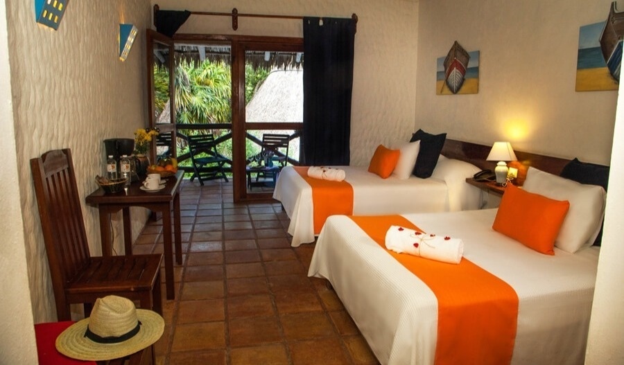 Una habitación de hotel con dos camas decoradas en blanco y naranja, una mesa con café y frutas, un balcón con vista a la vegetación y arte náutico en las paredes.