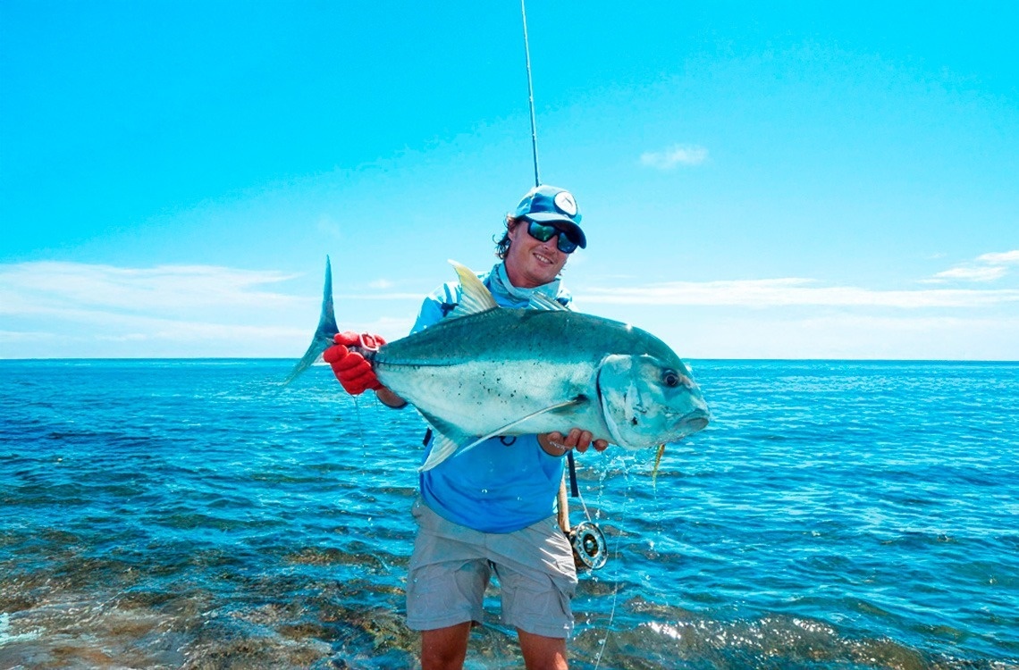 Un pescador sonriente sostiene un gran pez plateado en el agua, bajo un cielo azul claro.