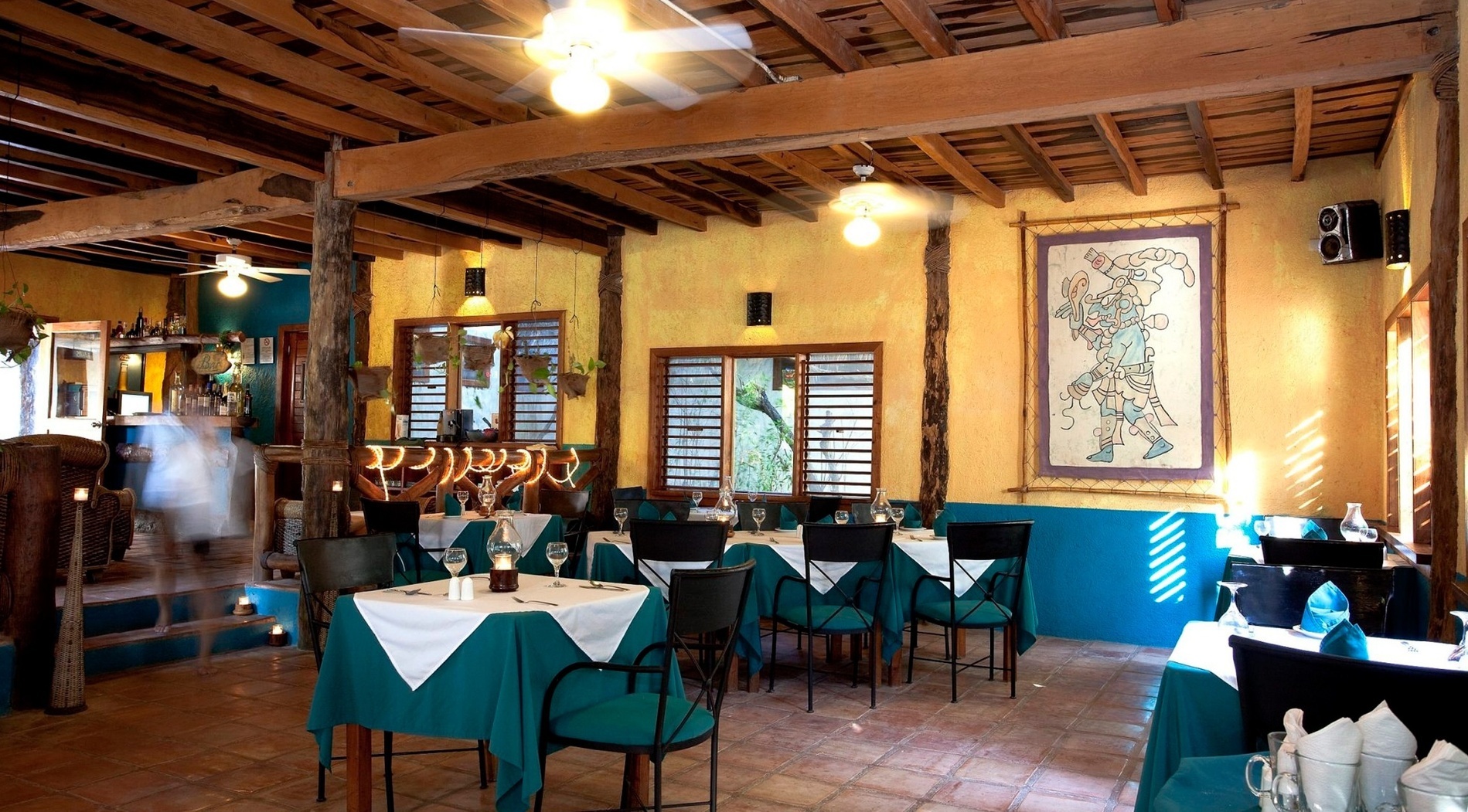 Un restaurante rústico de ambiente tropical con techos de vigas de madera, paredes amarillas y azules, mesas preparadas y un mural de temática maya.