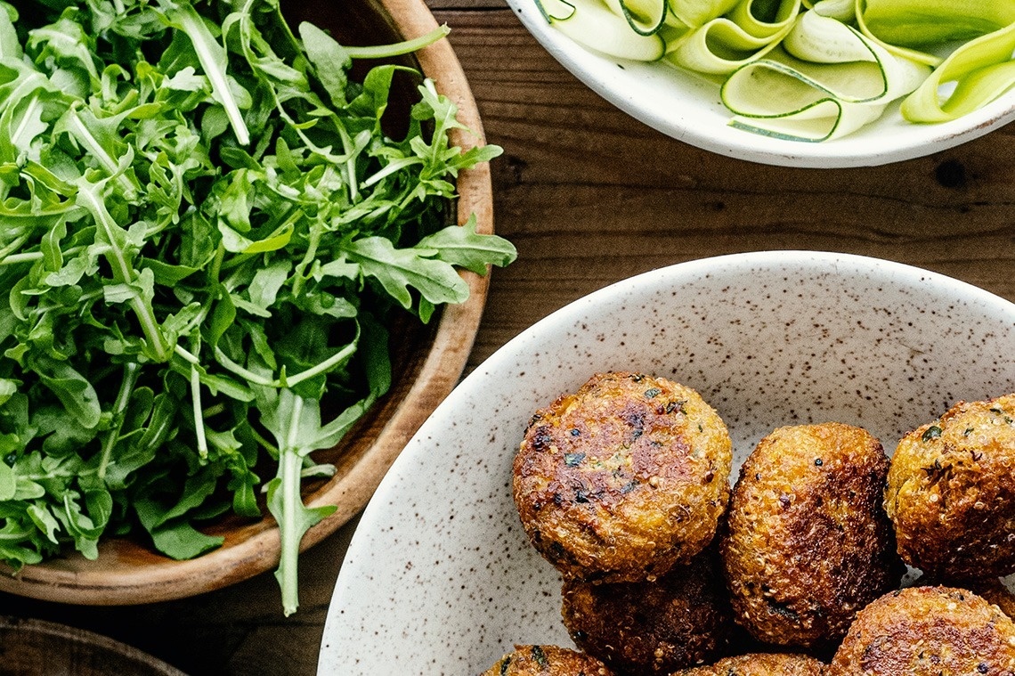 Una vista aérea de una mesa de madera presenta tres tazones: uno lleno de rúcula fresca, otro con cintas de pepino y un tercero con croquetas doradas.