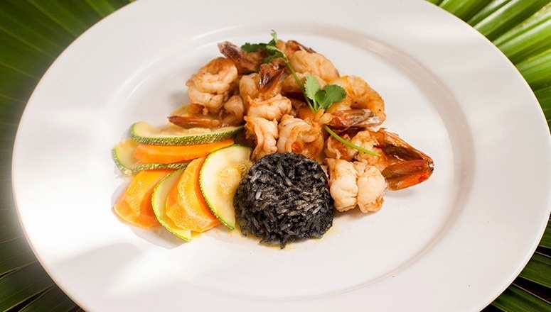 En un plato blanco se presenta un platillo de camarones salteados, arroz negro y vegetales en rodajas, sobre un fondo de hoja de palma verde.