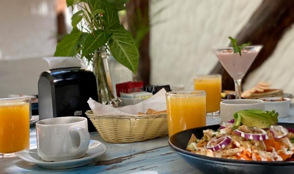 Un desayuno o brunch completo con jugo de naranja, café, un batido, una ensalada con aguacate y una cesta de pan, dispuesto sobre una mesa de madera.